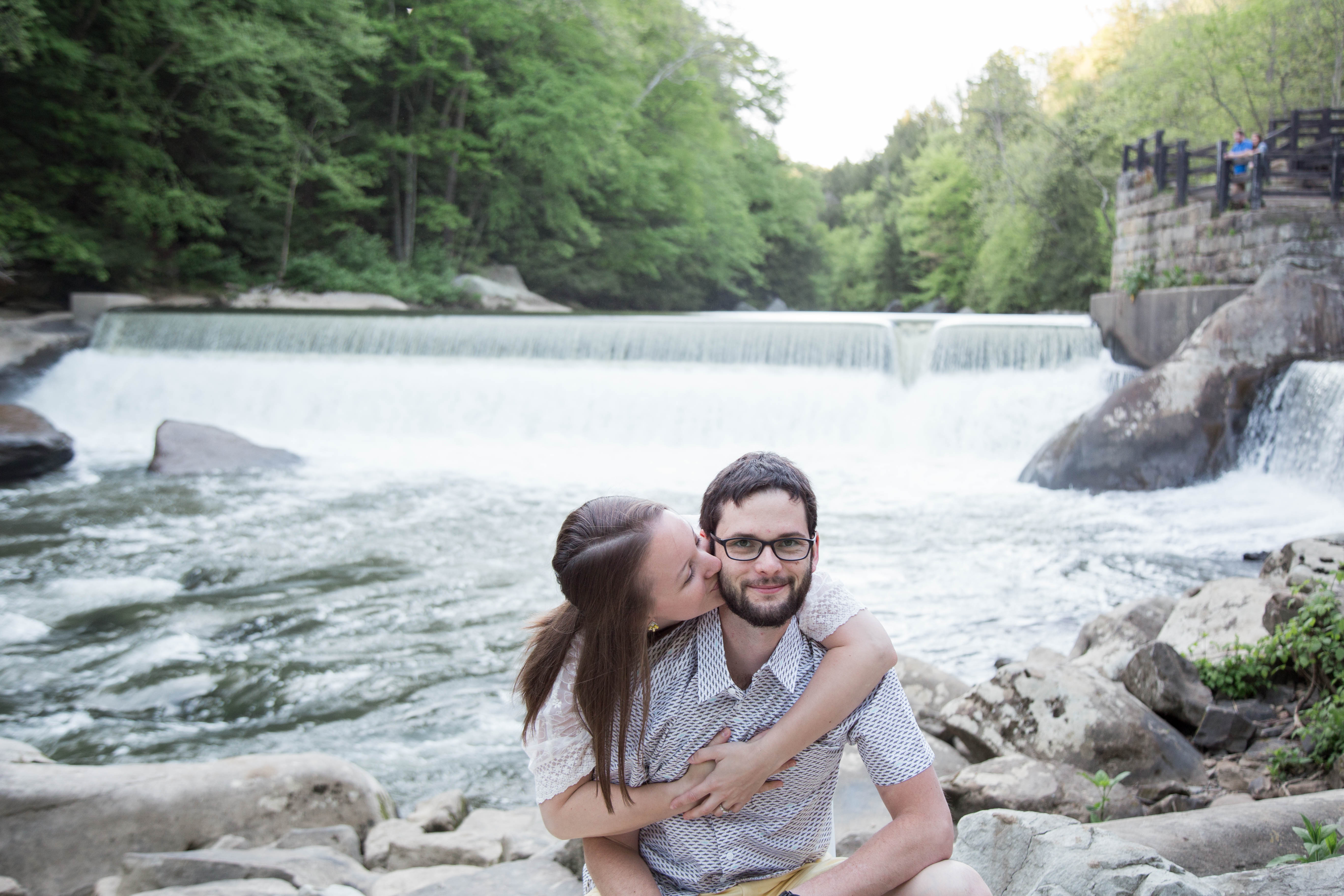 man kneels on rock while his fiancee wraps her arms around him and kisses his cheek