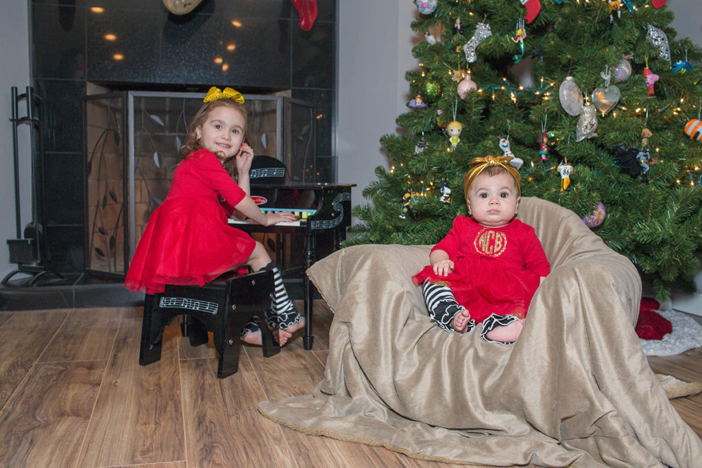 older sister sits at toy piano as her baby sister sits in a chair in front of the chirstmas tree