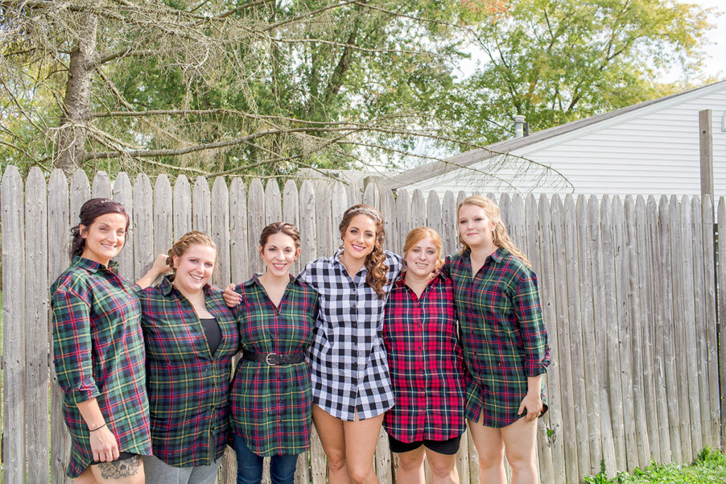 bride and bridesmaids wear flannel shirts and stand in front of fence outside of salon