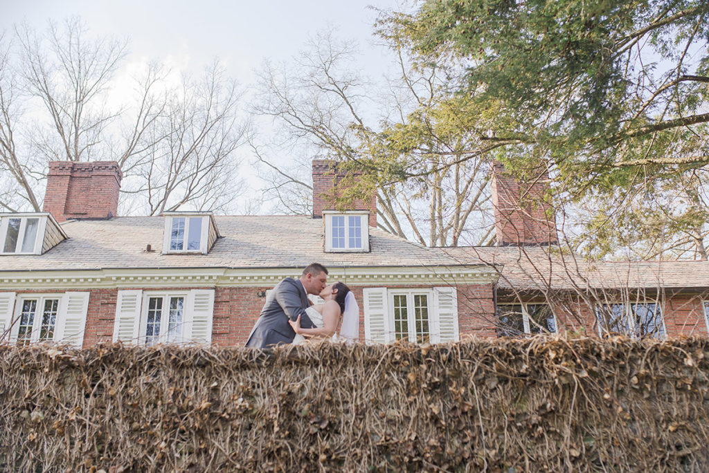 bride and groom stand at top of stone wall in front of mansion at robin hill park 