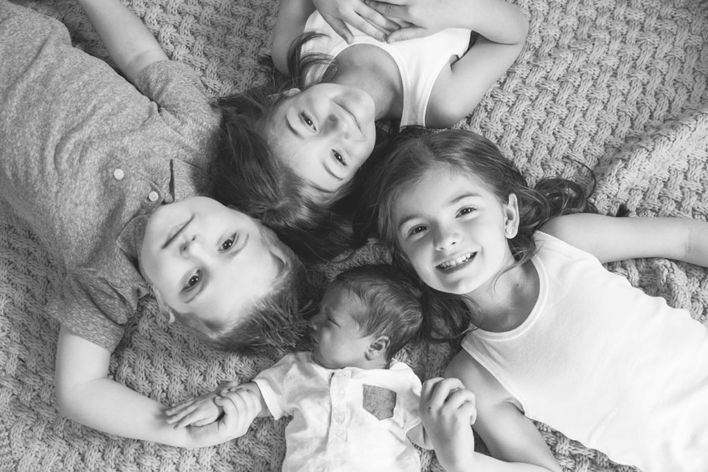 three siblings lie on their backs on the floor with their new baby brother