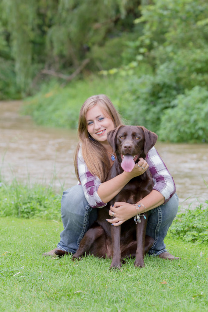 senior girl and her chocolate lab, maggie, sit in front of creek in milesburg
