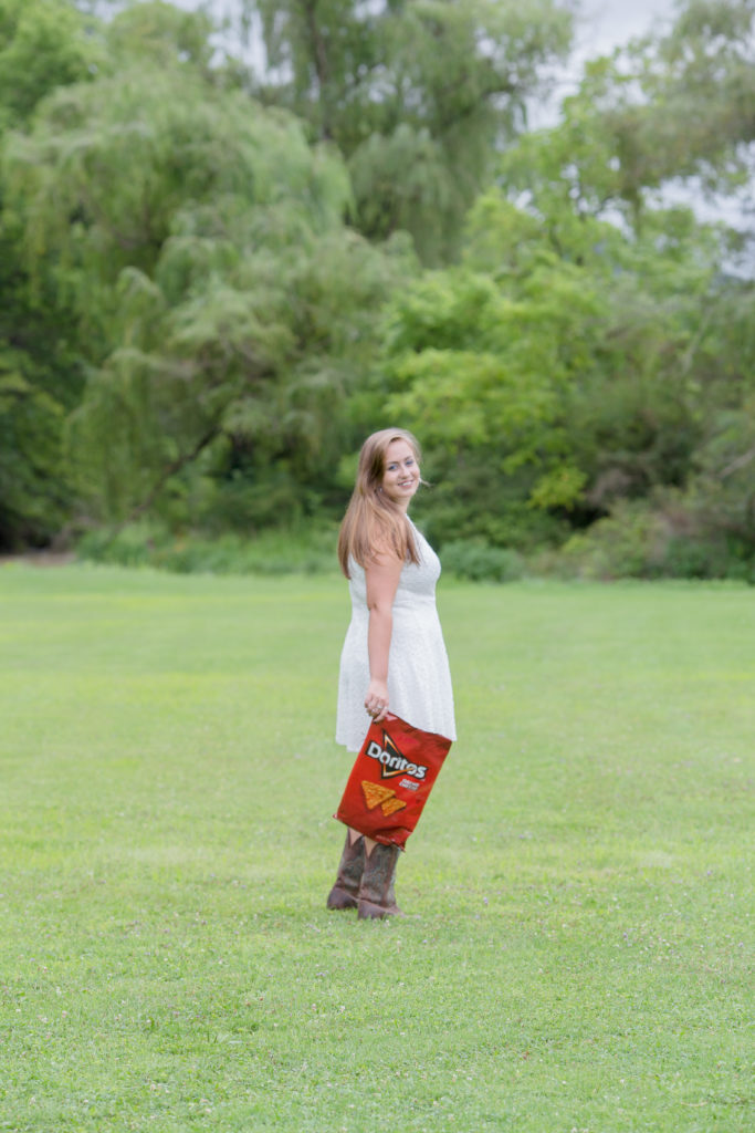 senior girl in white dress and cowboy boots stand on large lawn at a residence in milesburg while holding a doritos bag