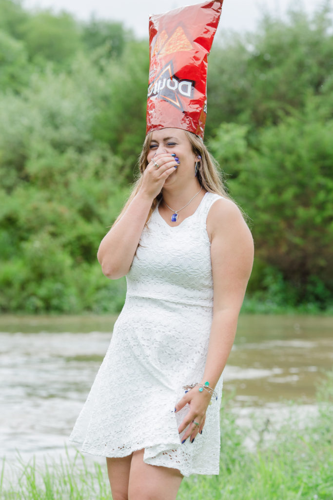 senior girl in white dress with doritos bag on her head stands in front of creek and laughs